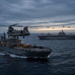 A CH-47 Chinook approaching the deck of a military support vessel at sea, with an amphibious ship, escorts, and a flight of helicopters headed toward a distant coastline